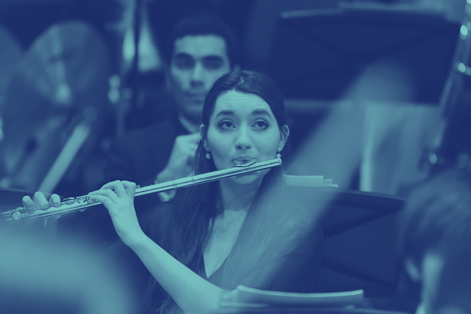 A blue filter over a flautist, wearing smart clothes, performing in an orchestra, facing the conductor.